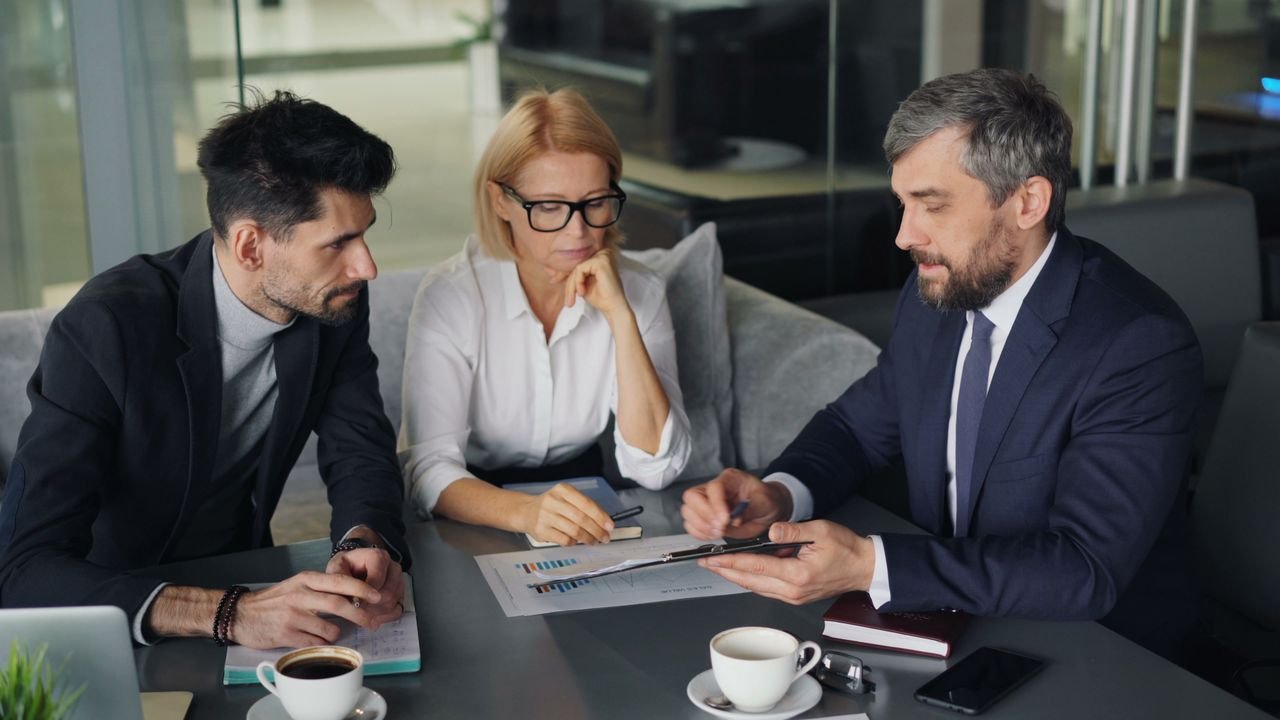 Three professionals engaged in a business discussion at an office table with coffee.